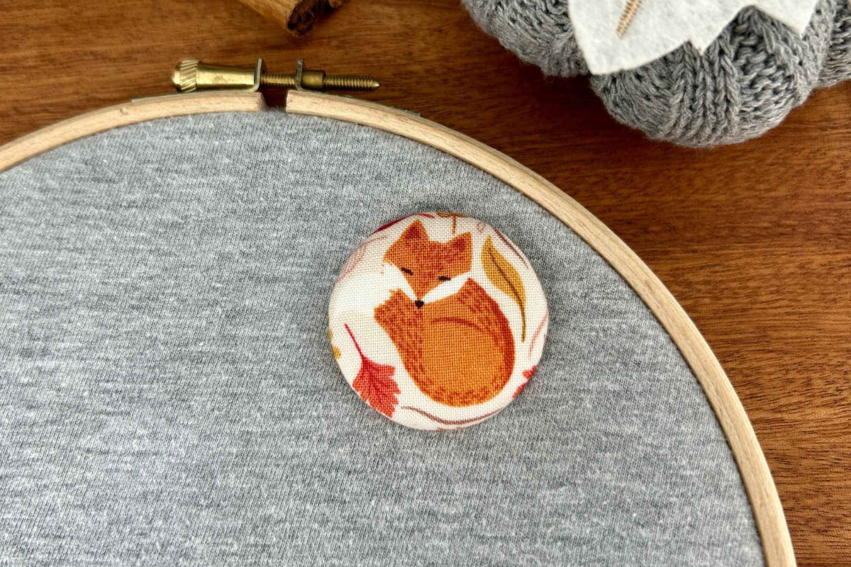 A round fabric magnetic needle minder featuring a hibernating fox design in orange, brown, and white, displayed on gray fabric inside a wooden embroidery hoop on a wooden table
