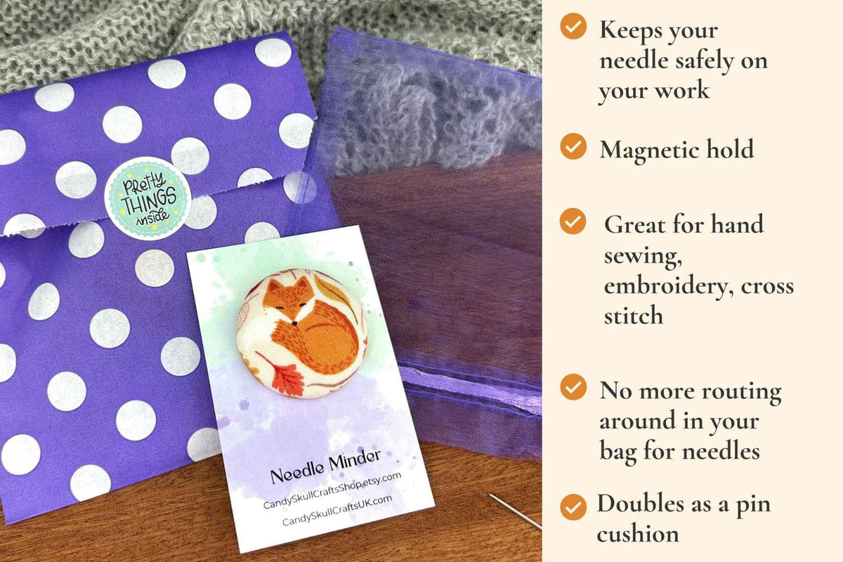 A product listing graphic showing a purple polka dot envelope, a magnetic needle minder featuring a hibernating fox design, and a branded card for 'needle Minder' on a wooden surface next to a text panel describing features. keeps your needle safely on your work. Magnetic hold great for hand sewing, embroidery, cross stitch. No more routing around in your bag looking for needles. Doubles as a pin cushion.
