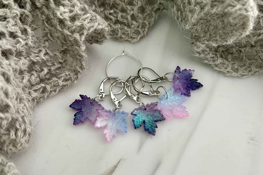5 knitting or crochet stitch markers with maple leaf charms in a purple, blue, and pink gradient, displayed on a white marble surface with a gray knitted scarf.