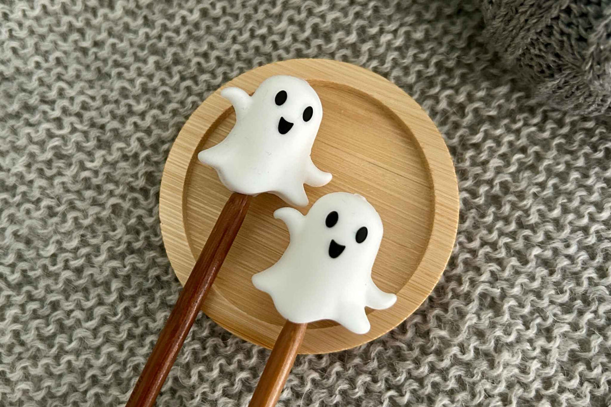 Two white ghost-shaped knitting needle toppers with black eyes and mouths rest on a round light wood plate over a gray knitted fabric background.