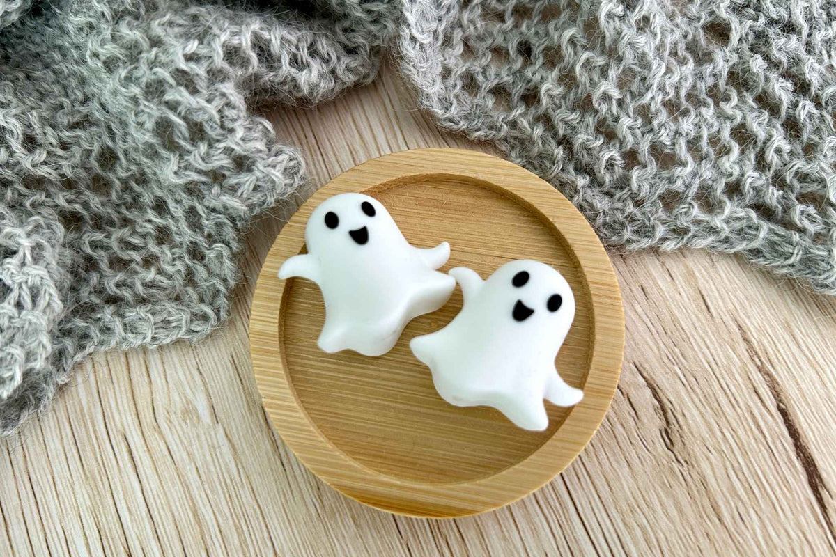 Two small white ghost-shaped stitch savers with black facial features rest on a round wooden tray, surrounded by gray knitted fabric on a light wood surface.