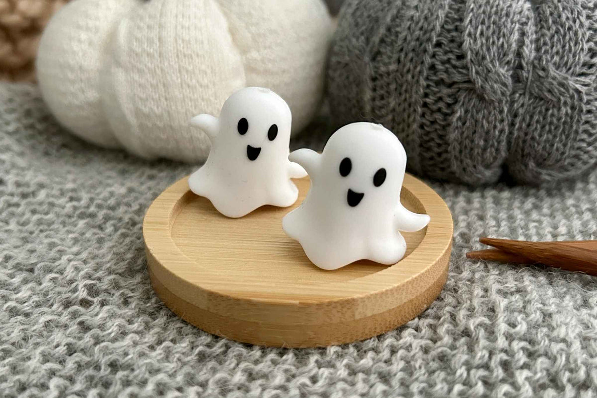 Two small white ghost-shaped knitting needle stoppers with black facial features sit on a round light wood tray, styled on a gray knitted surface with soft knit props in the background.