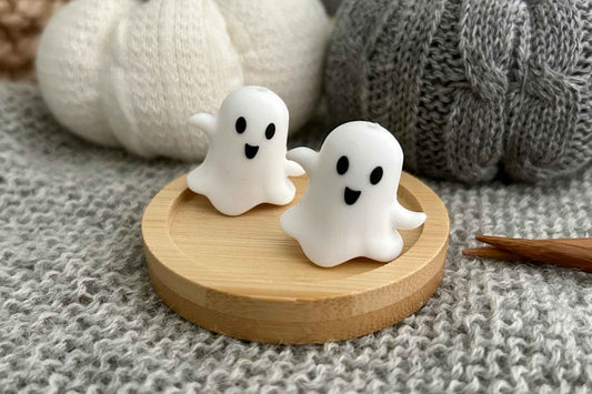 Two small white ghost-shaped knitting needle stoppers with black facial features sit on a round light wood tray, styled on a gray knitted surface with soft knit props in the background.