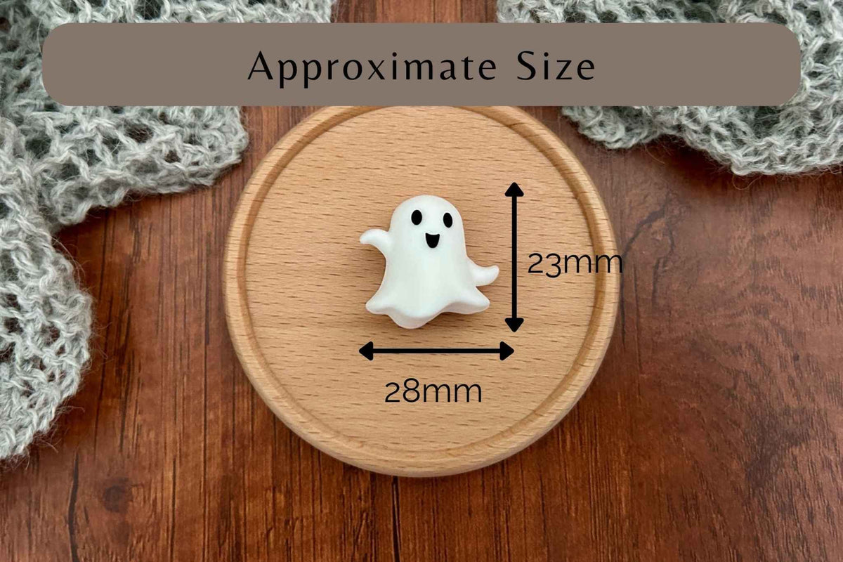 A small white ghost-shaped knitting accessory displayed on a round wooden coaster over a wooden surface, with overlaid arrows and text showing approximate dimensions of 28mm by 23mm.