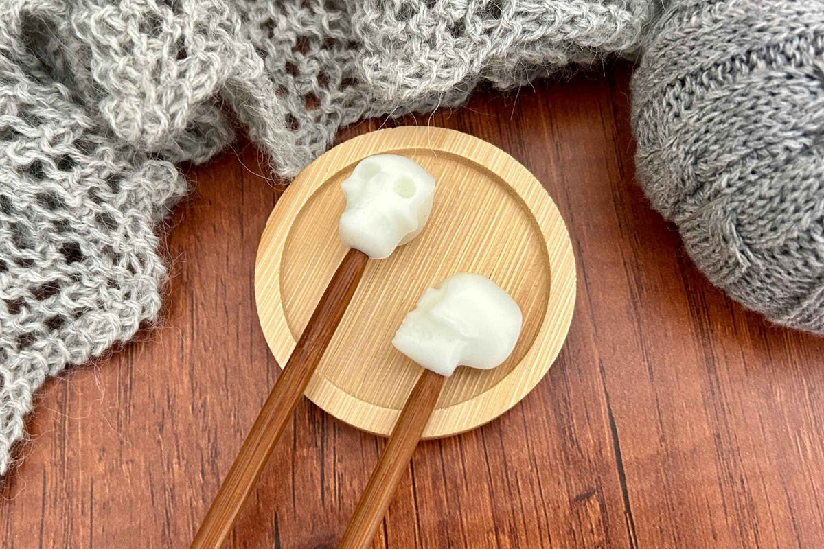 Two skull-shaped knitting needle stoppers on wooden needles resting on a round bamboo plate, with gray knitted yarn and a gray knit scarf on a wooden tabletop.
