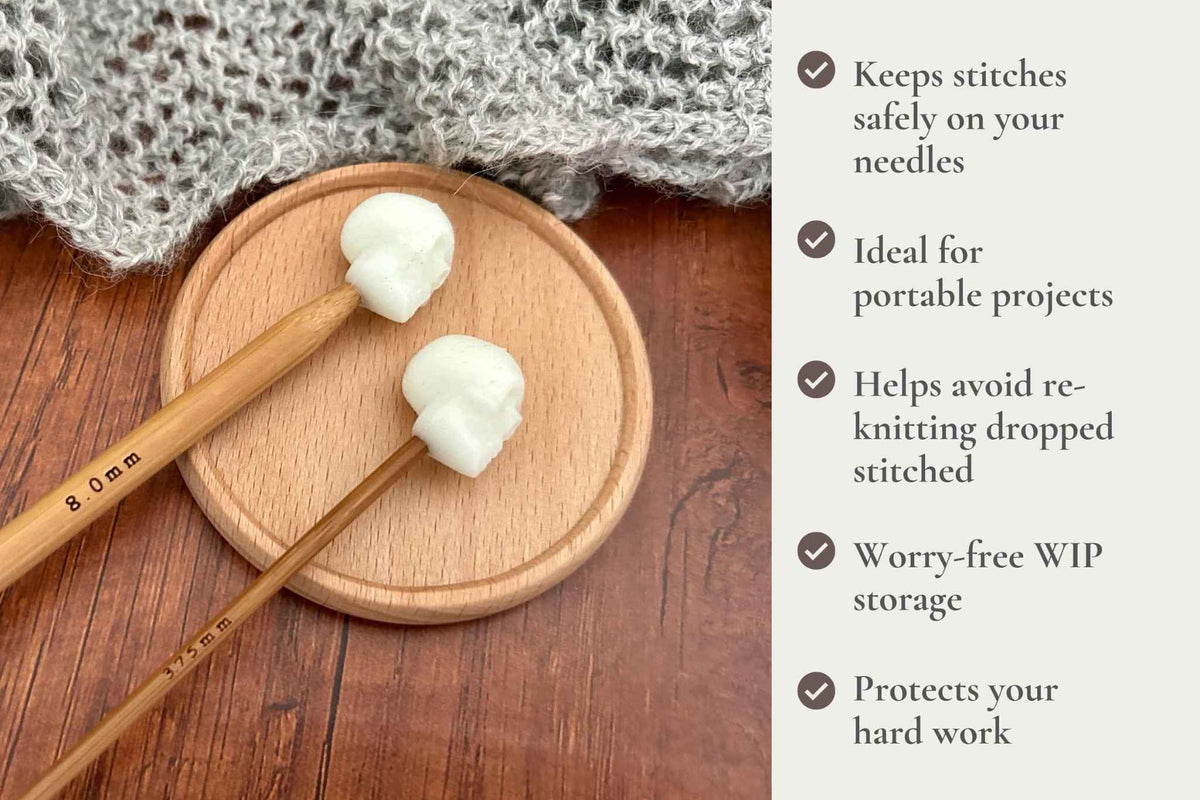 Two wooden knitting needles with 3D skull-shaped stitch protectors rest on a round wooden tray, with a knitted fabric nearby and a text panel on the right describing benefits. Keeps stitches on needles. Ideal for portable projects. Helps avoid re-knitting dropped stitches. Fits mm to 2.5mm to 8mm needles 

