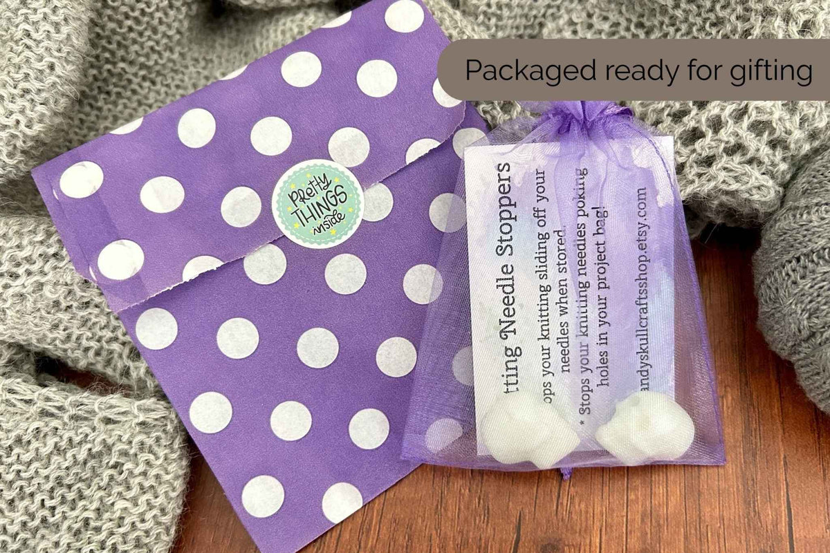 Purple and white polka dot gift packaging with a small organza bag containing skull knitting needle stoppers, and an instruction card, styled on a wooden surface with a gray knit blanket. A text overlay reads 'Packaged ready for giving'.