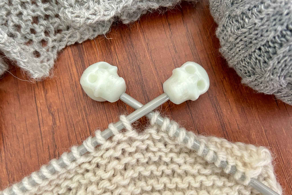 Close-up of knitting needles with skull-shaped stoppers on the tips, shown in use with cream and gray yarn on a wooden surface.