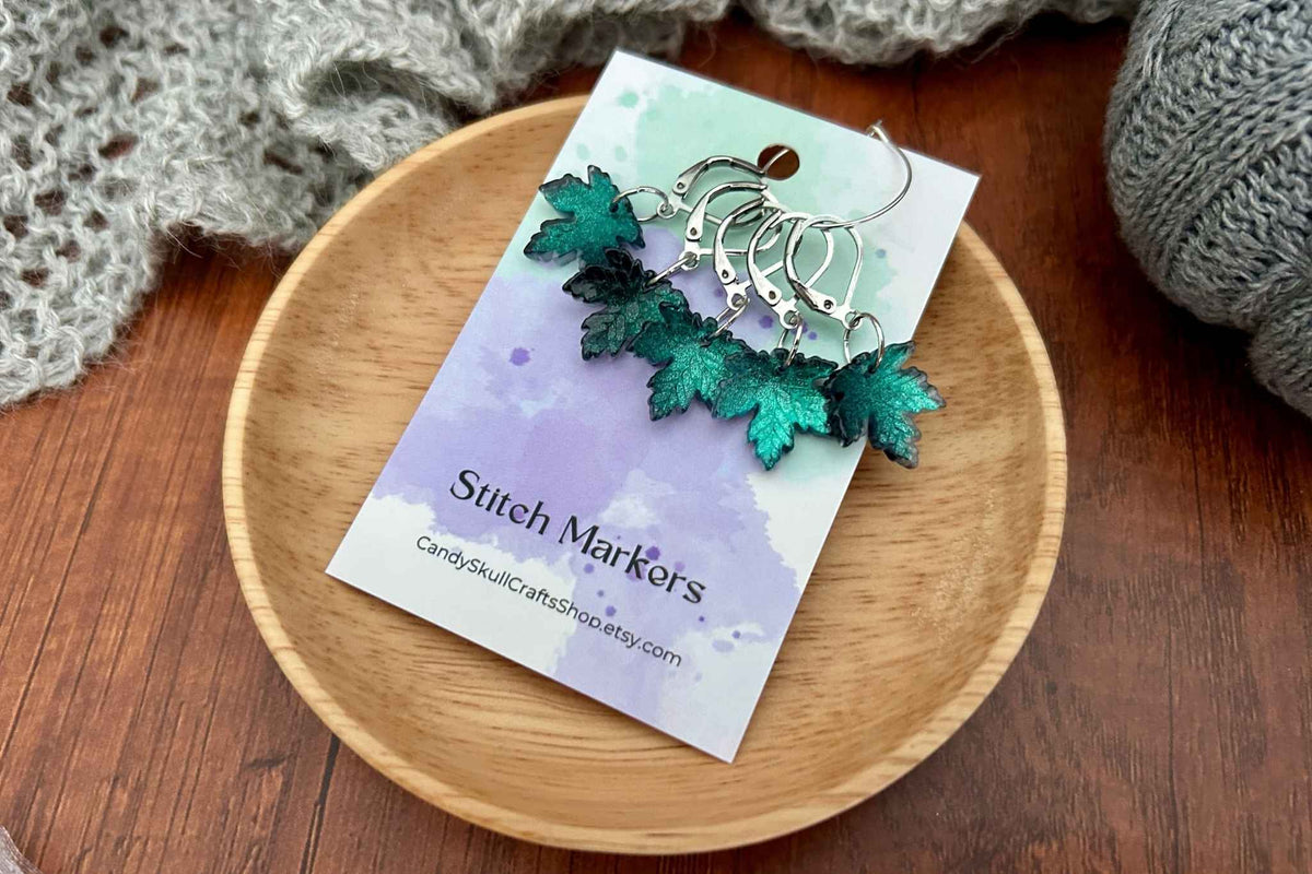 A set of knitting stitch markers with glittery teal and green maple leaf charms, displayed on a branded card labelled 'Stitch Markers' resting on a small wooden dish over a wood table.
