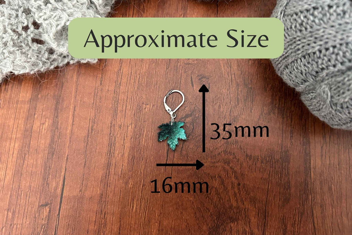 A small maple leaf-shaped knitting or crochet stitch marker shown on a wooden surface with overlaid arrows and text indicating approximate dimensions of 16mm by 35mm. Knitted gray fabric appears at the top corners, and a green label at the top reads 'Approximate Size'.