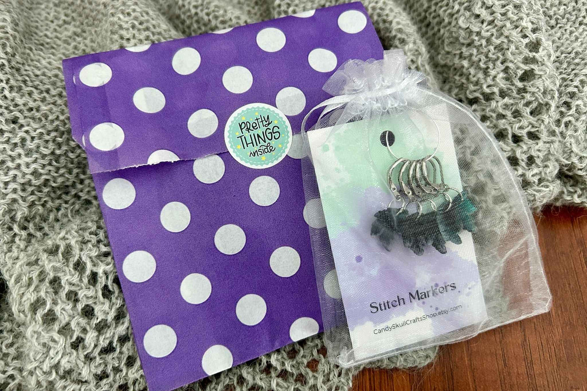 A purple and white polka dot envelope on a gray knitted blanket next to an organza bag with 5 knitting stitch markers on a branded card