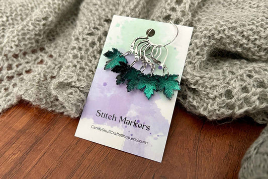 A set of 5 black and green gothic maple leaf knitting stitch markers, displayed on a printed card labelled 'Stitch Markers' and placed on a wooden surface with a gray knitted fabric in the background.