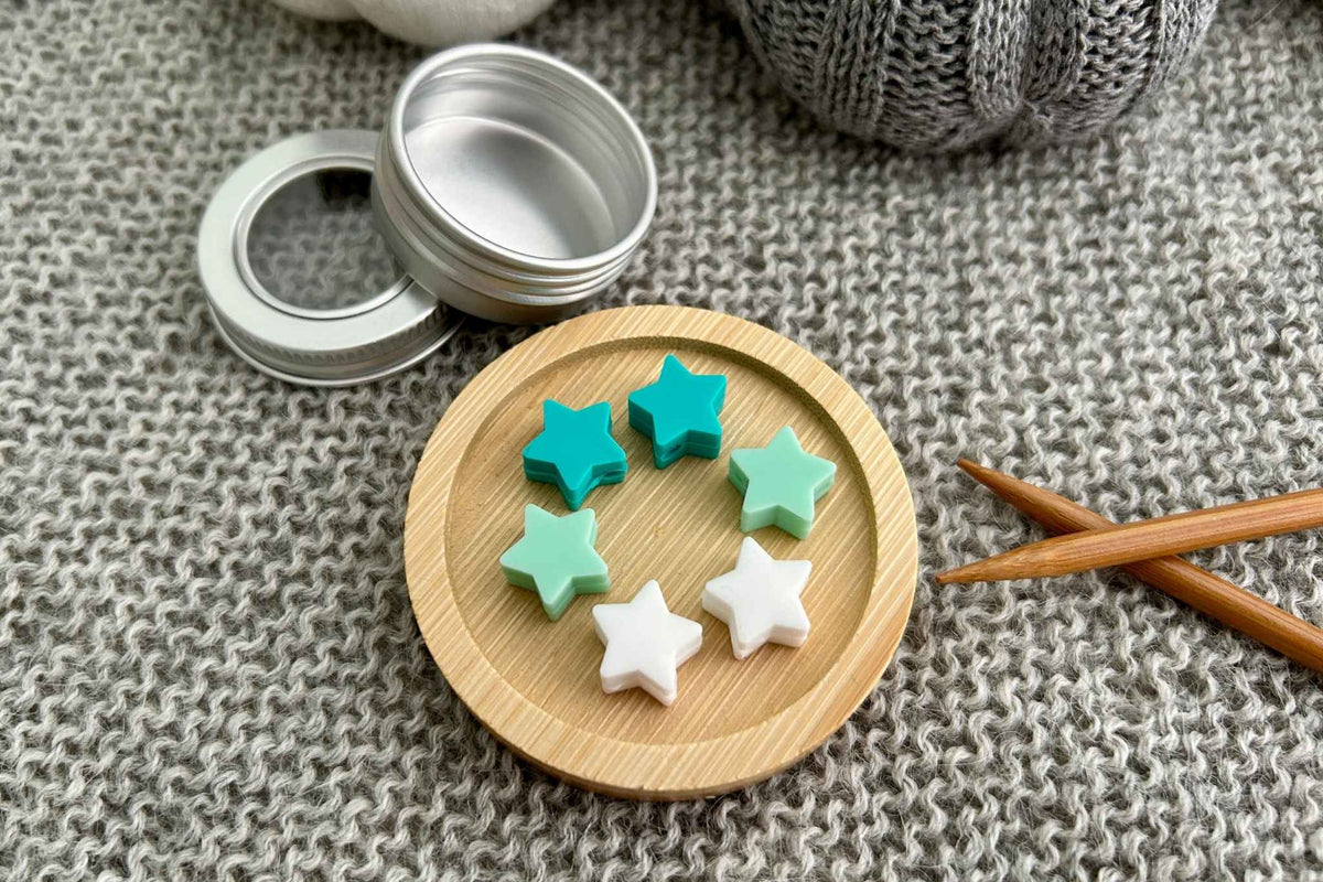 Five small star-shaped knitting needle stitch stoppers in mint green, teal, white, and light pink arranged on a round wooden tray, with an open silver metal tin nearby on a gray knitted fabric background.