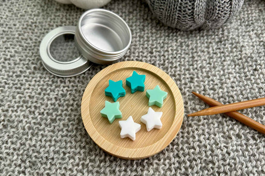 Five small star-shaped knitting needle stitch stoppers in mint green, teal, white, and light pink arranged on a round wooden tray, with an open silver metal tin nearby on a gray knitted fabric background.
