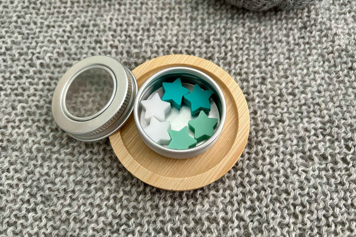 A small round metal tin with a silver lid resting beside it on a light wood coaster, filled with star-shaped knitting needle stoppers in white, mint green, and teal, set on a gray knitted blanket.