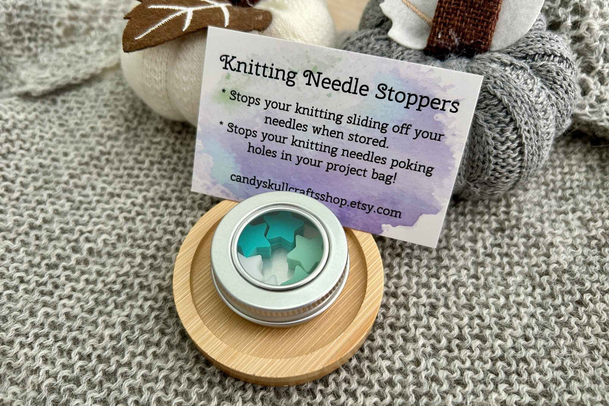 A small round tin of knitting needle stoppers sits on a wooden coaster on a knitted blanket, with a printed card behind it reading 'Knitting Needle Stoppers' and 'candyskullercraftsshop.etsy.