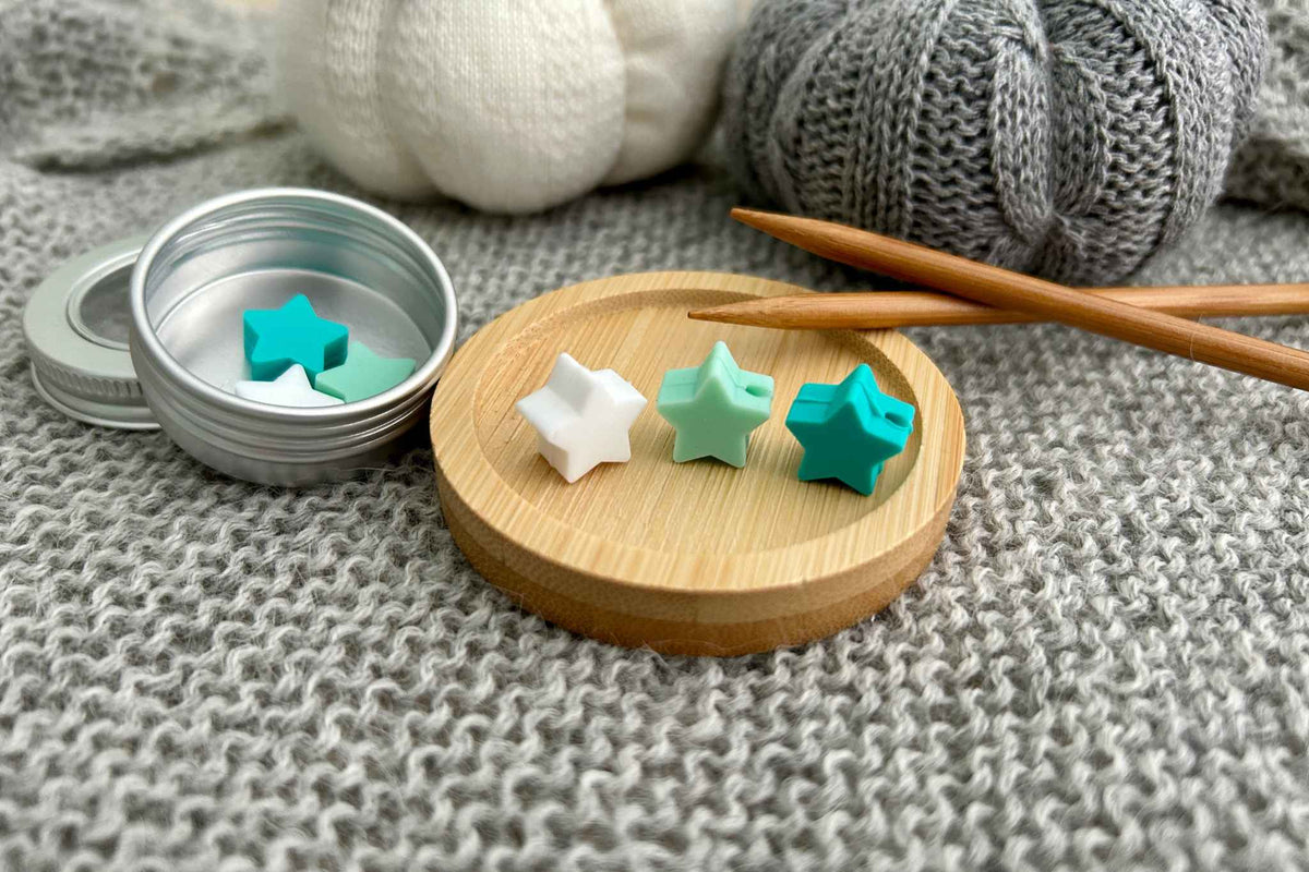 Three small star-shaped silicone knitting needle stoppers in white, mint green, and teal are displayed on a round wooden tray next to an open metal tin, with wooden knitting needles and knitted yarn in the background.