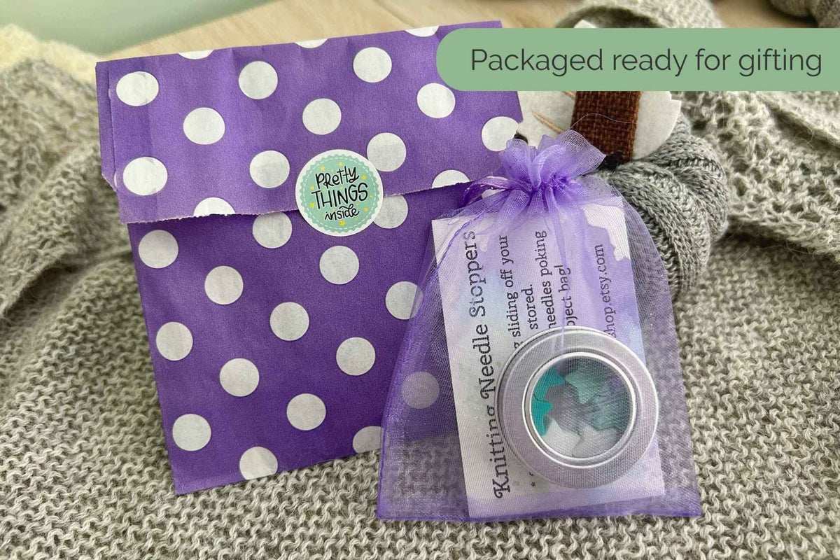 A small round tin of knitting needle stoppers in a clear mesh gift bag tied with a purple ribbon, placed in front of a purple polka dot envelope on a knitted blanket. A green banner reads 'Packaged ready for giving'.