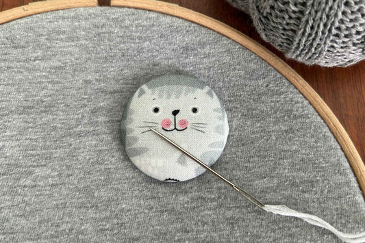 A small round fabric needle minder featuring a white and gray cat face with pink cheeks, shown on gray fabric inside a wooden embroidery hoop with a needle and yarn visible.