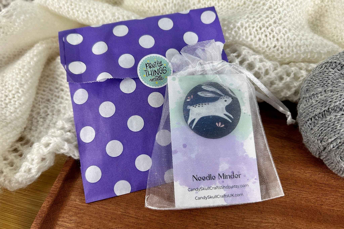 A white rabbit or hare fabric magnetic needle minder in a white mesh organza bag. Shown next to the packaging of a purple an white polka dot bag with a stickers which reads "pretty things inside"