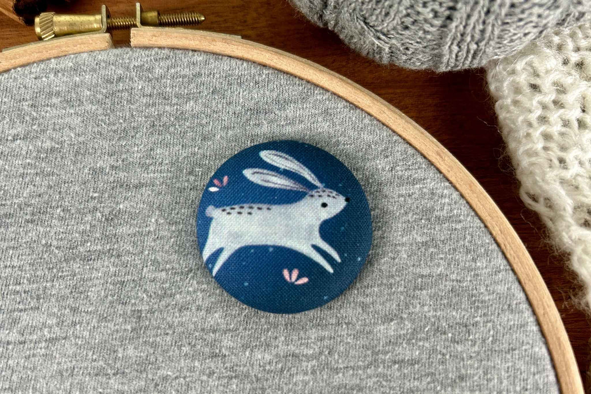 A round fabric magnetic needle minder featuring a white rabbit /hare on a dark blue background, displayed on a gray fabric hoop with knitting materials nearby.