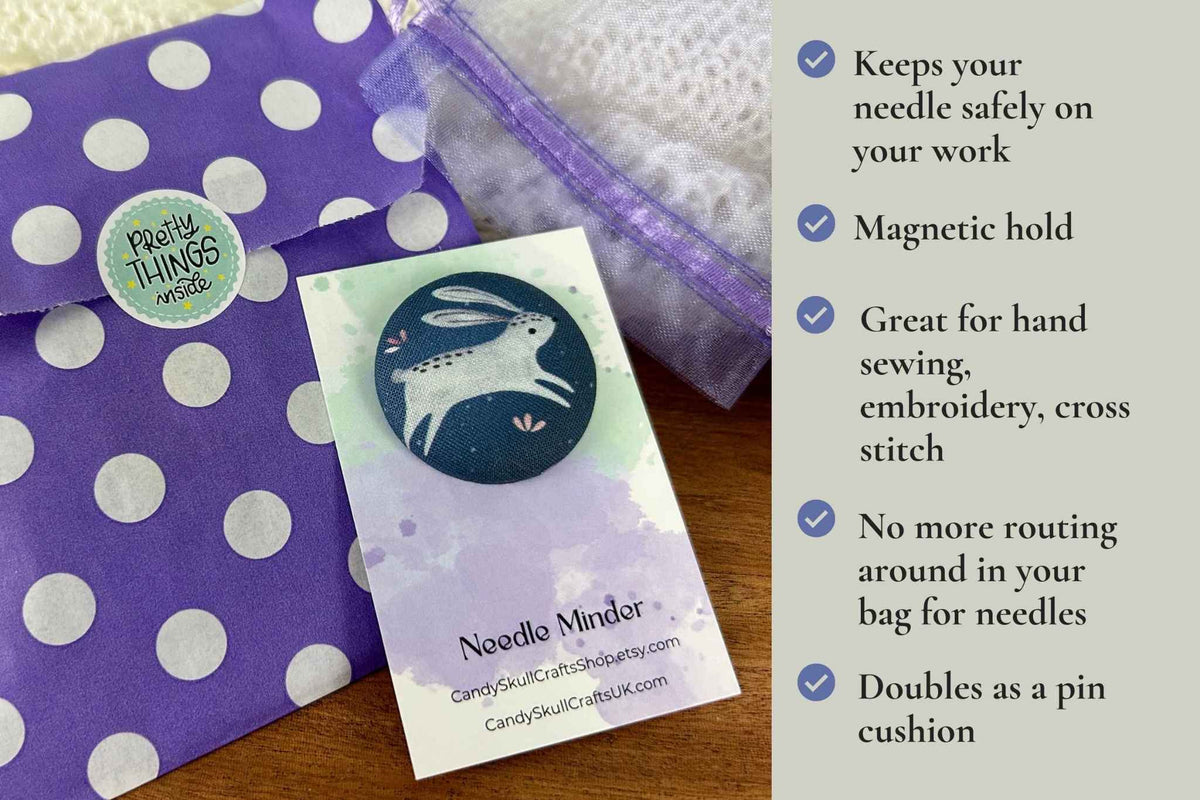 a fabric magnetic needle minder featuring a white rabbit on a dark blue background next to it's packaging: a mesh organza bag and a polka dot paper bag. The benefits are listed as:  keeps your needle safely on your work. Magnetic hold great for hand sewing, embroidery, cross stitch. No more routing around in your bag looking for needles. Doubles as a pin cushion.
