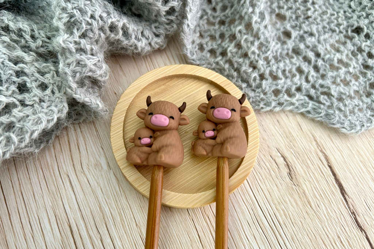 two highland cow shaped needle stitch savers on wooden needles displayed on a small round wooden tray over a brown wood surface with a gray knitted textile in the background. The mummy cow is cuddling her little calf.
