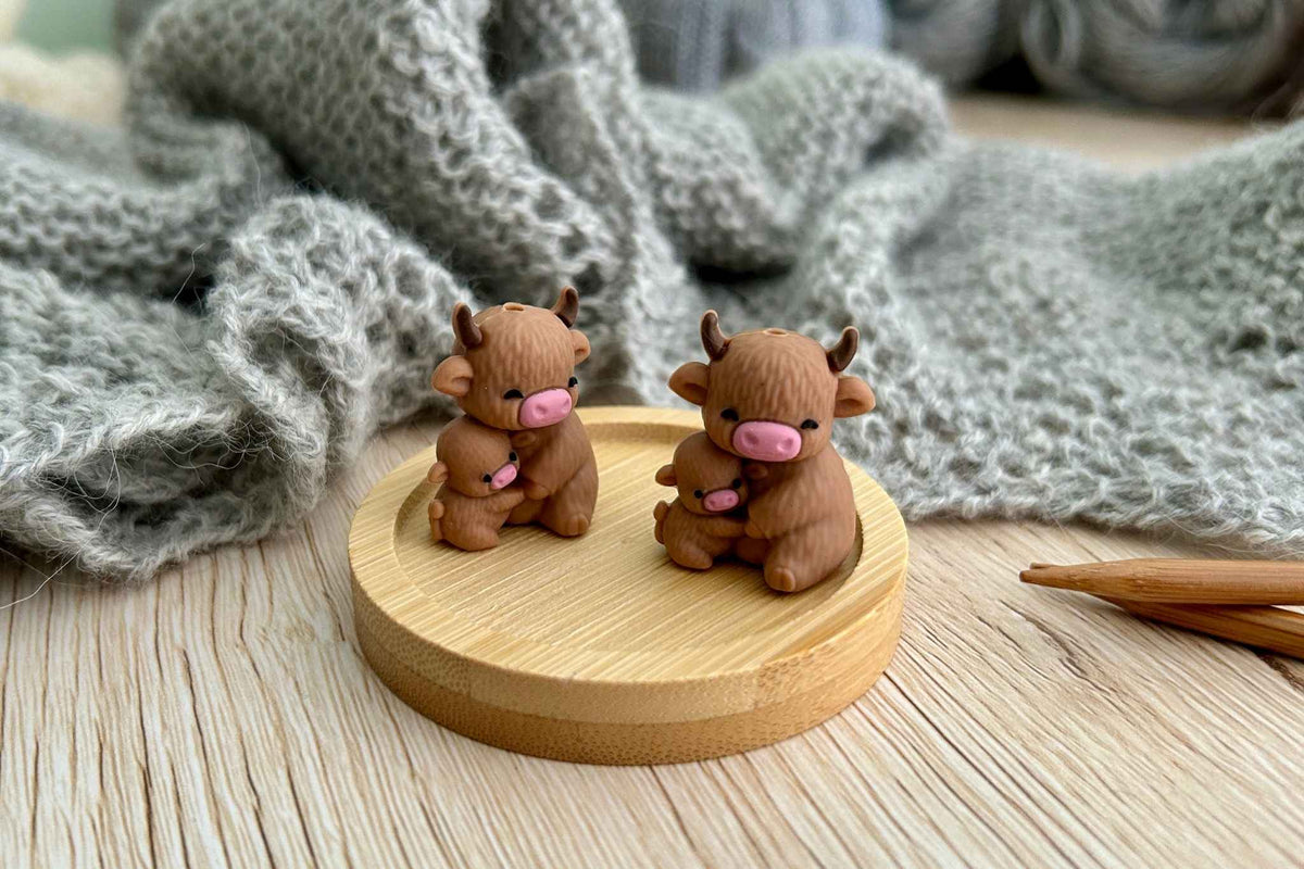 A close up of the highland cow stitch savers with a gray knitted blanket and wooden knitting needles in the background.