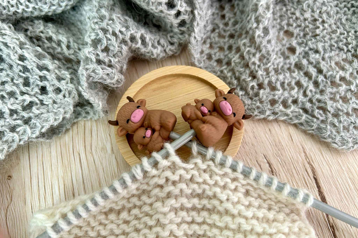 Two Aberdeen Angus highland cow knitting needle stoppers stopping stitches falling off cream knitting, with a gray knitted fabric in the background on a wooden surface.

