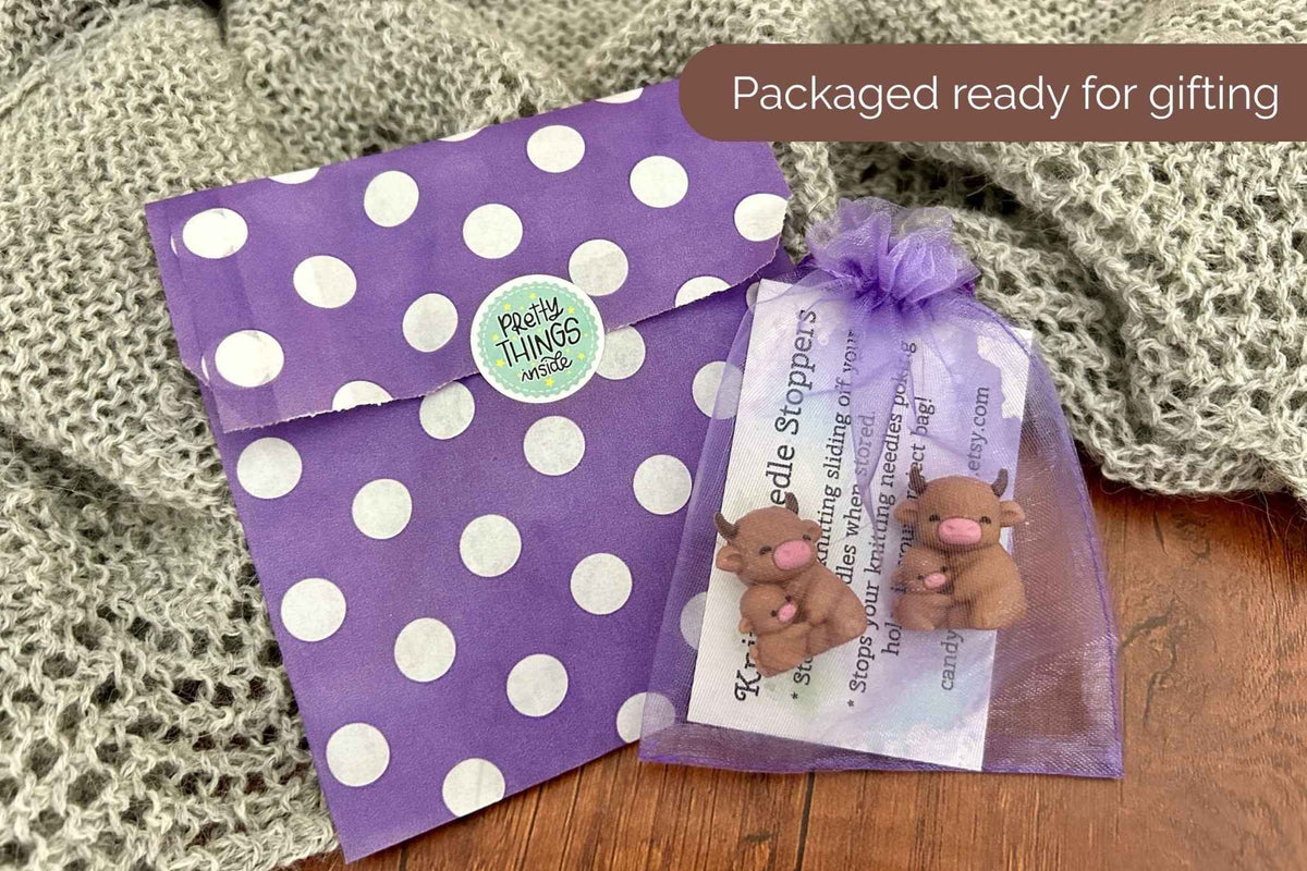 A gift set featuring a purple and white polka dot envelope and a sheer purple organza bag with a set of 2 needle stoppers and an instruction card, displayed on a wooden surface with a knitted blanket.