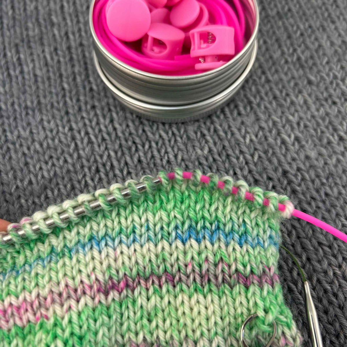 A close-up of a knitted fabric swatch with green, pink, and blue stripes on knitting needle being slid onto a pink knitting stitch holder cord, with a small metal tin of bright pink cords and stoppers in background on a gray knitted surface