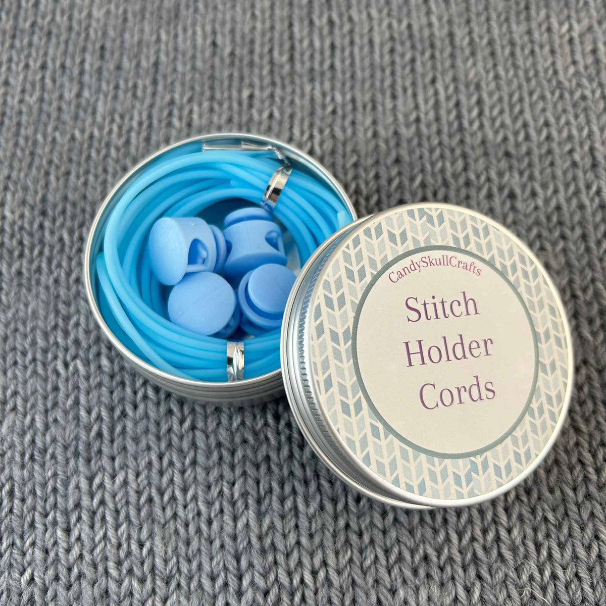 Blue knitting stitch holder cords and 4 stoppers in a round metal tin, with the lid labelled 'Candy Skull Crafts Stitch Holder Cords' resting beside the open tin on a gray knitted fabric background.