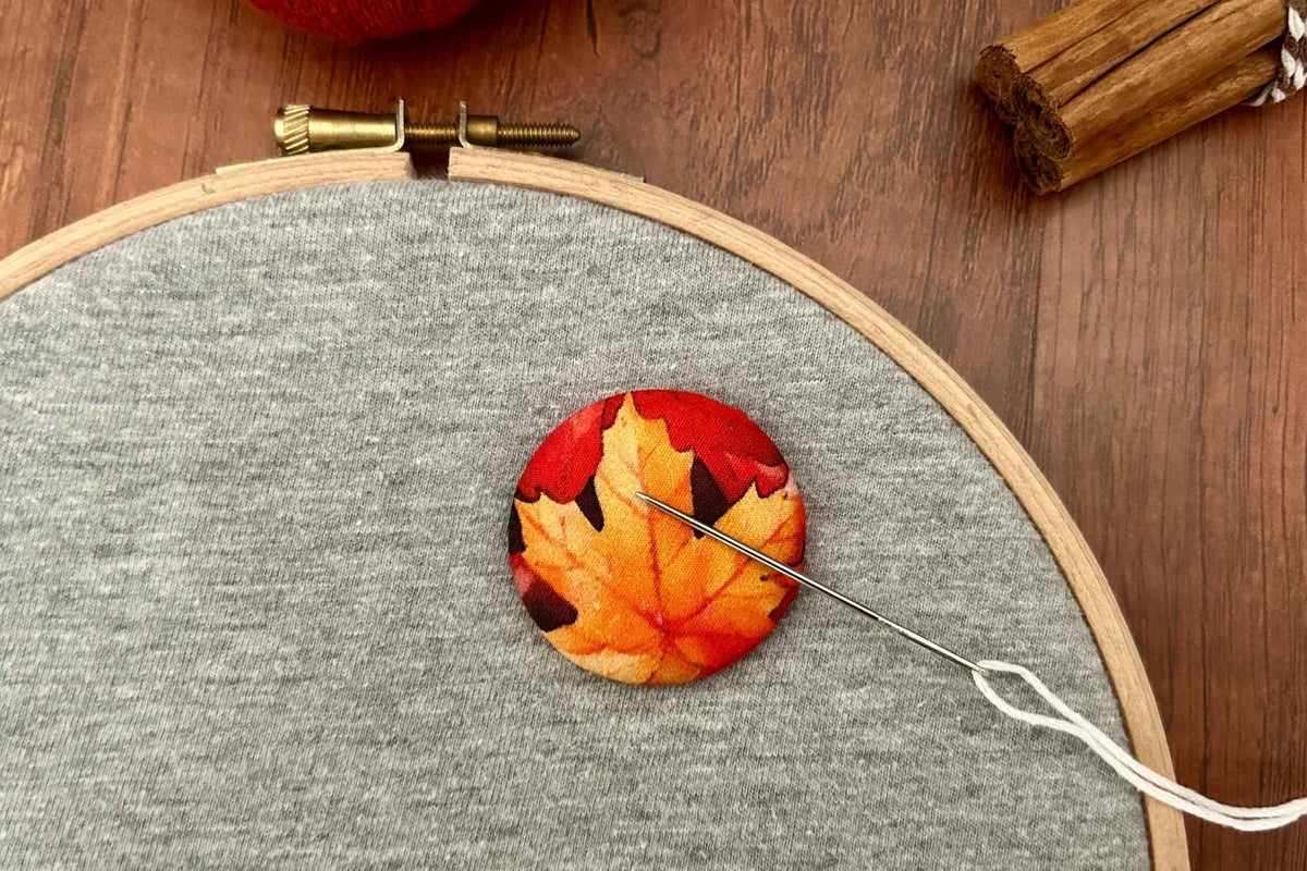 A close-up of a magnetic needle minder  featuring a printed autumn maple leaf pattern in red, orange, and yellow, sewn onto gray fabric inside a wooden embroidery hoop. A needle and thread are visible, with a wooden surface and cinnamon sticks in the background.