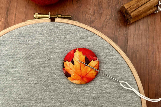 A close-up of a magnetic needle minder  featuring a printed autumn maple leaf pattern in red, orange, and yellow, sewn onto gray fabric inside a wooden embroidery hoop. A needle and thread are visible, with a wooden surface and cinnamon sticks in the background.