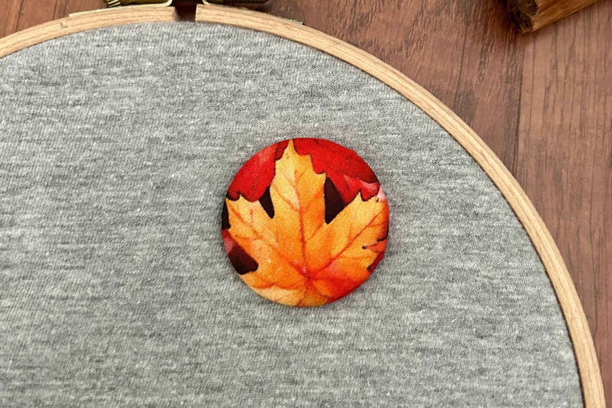 A circular fabric magnetic needle minder  featuring a printed autumn maple leaf in shades of orange, yellow, and red, displayed on a gray heathered t-shirt inside a wooden embroidery hoop over a dark wood surface.