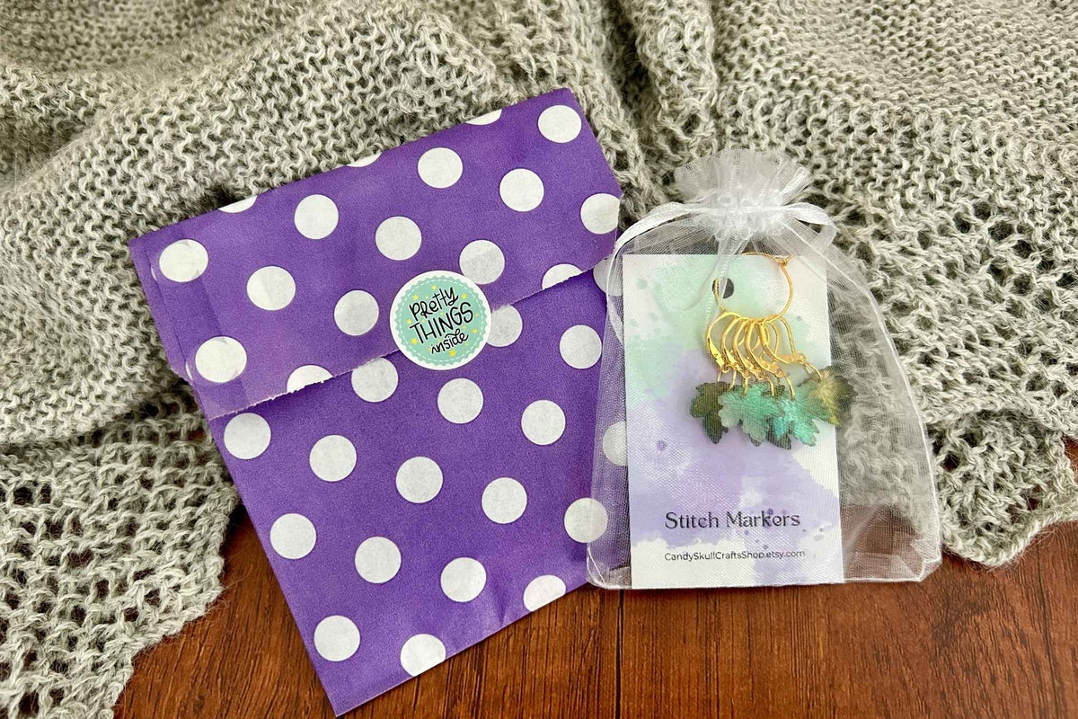 A purple paper gift bag with white polka dots and a round 'pretty things inside.' sticker, next to a small clear bag containing gold and green knitting or crochet maple leaf stitch markers on a card, set on a wooden surface with a knitted blanket in the background.