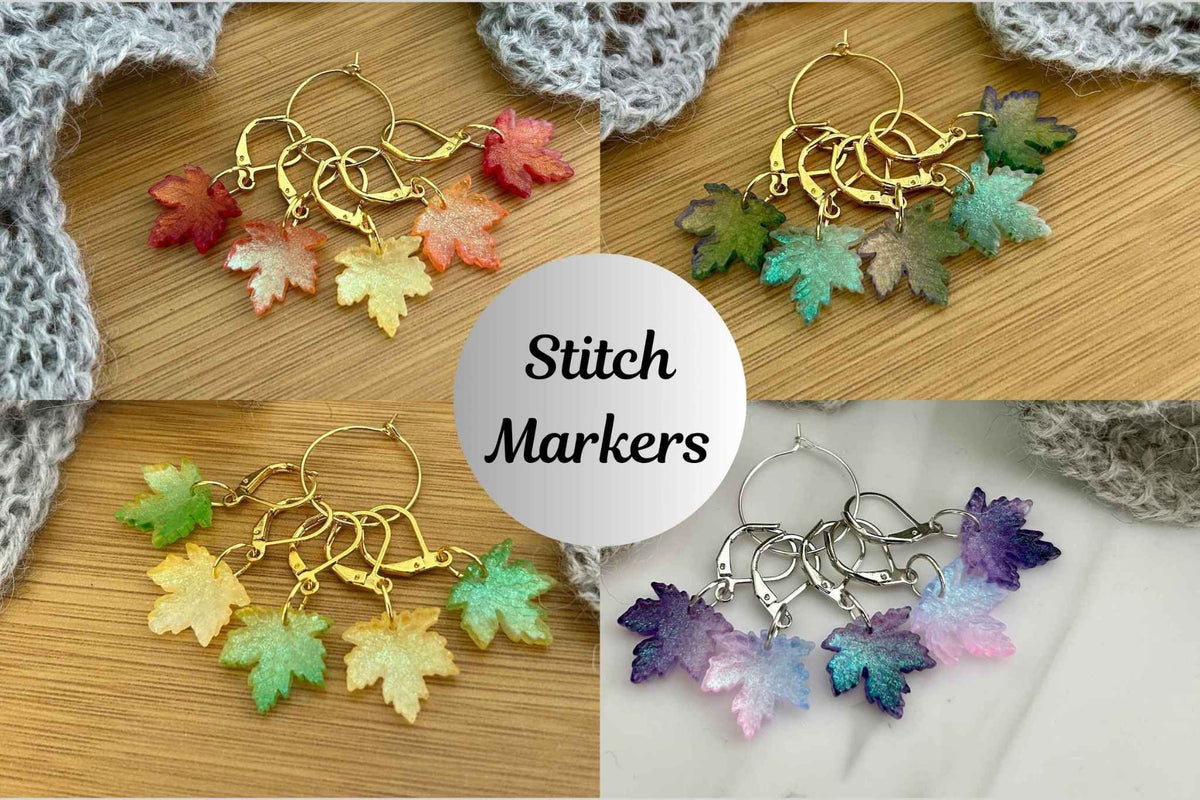 Four sets of maple leaf knitting and crochet stitch markers in 4 colourways to match the seasons, arranged on a wooden surface with a gray crocheted cloth. A central white circle reads 'Stitch Markers'.