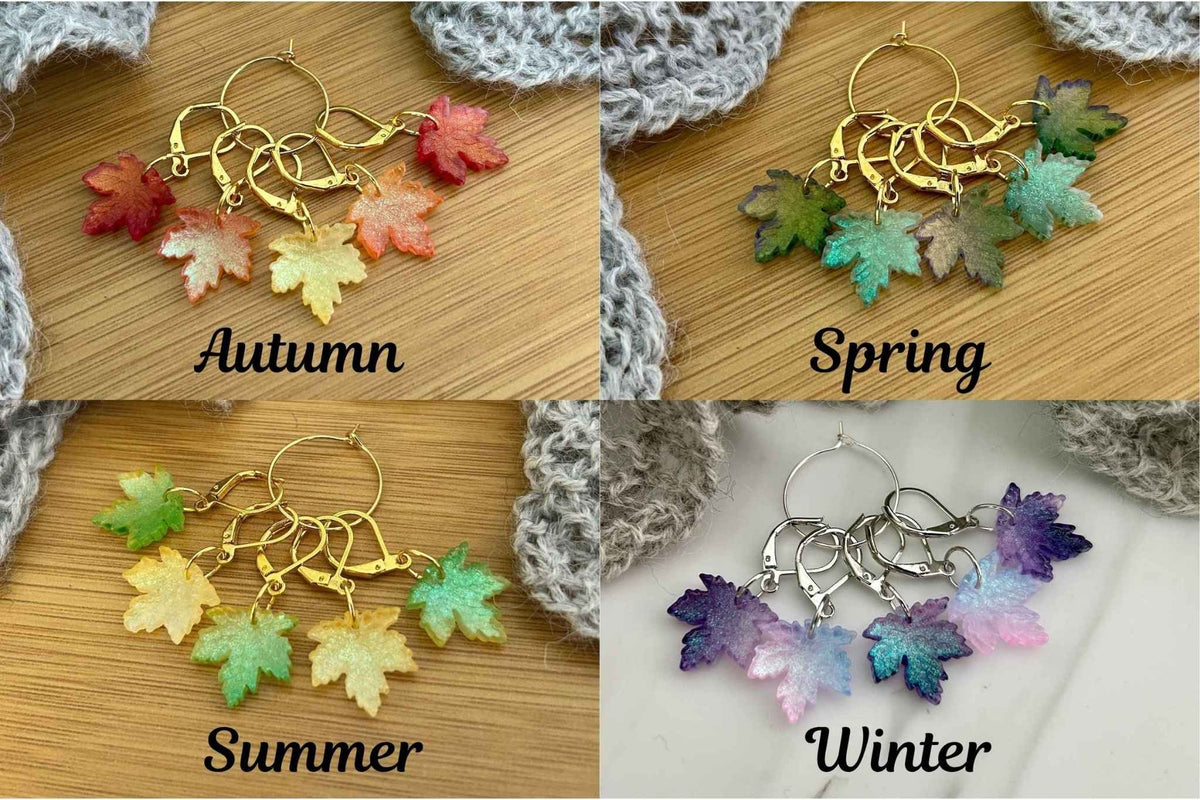 Four pairs of knitting and crochet stitch markers with glass autumn leaves, arranged in a 2x2 grid labelled Autumn, Spring, Summer, and Winter. The top-left pair has red, orange, and yellow leaves on gold hoops; the top-right has green and blue leaves on gold hoops; the bottom-left has green, yellow, and light green leaves on silver hoops; the bottom-right has purple, blue, and pink leaves on silver hoops.