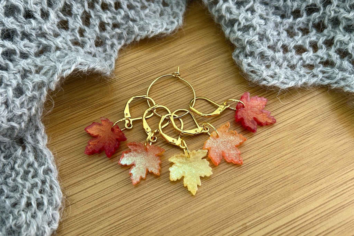 A set of 5 maple leaf knitting or crochet stitch markers or progress keepers with dangling maple leaf charms in red, orange, and yellow, displayed on a wooden surface with a gray knitted fabric in the background.