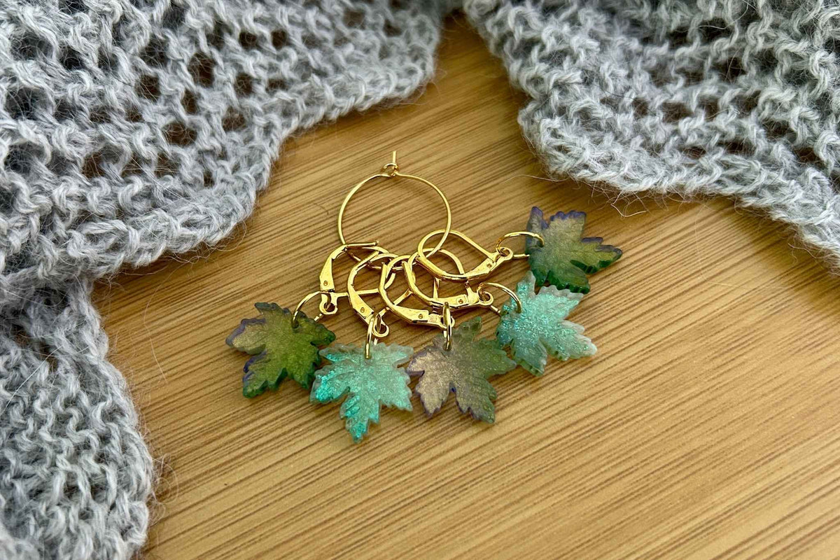 A set of 5 maple leaf knitting or crochet stitch markers or progress keepers with dangling maple leaf charms in various shades of green, displayed on a wooden surface with a gray knitted fabric in the background.