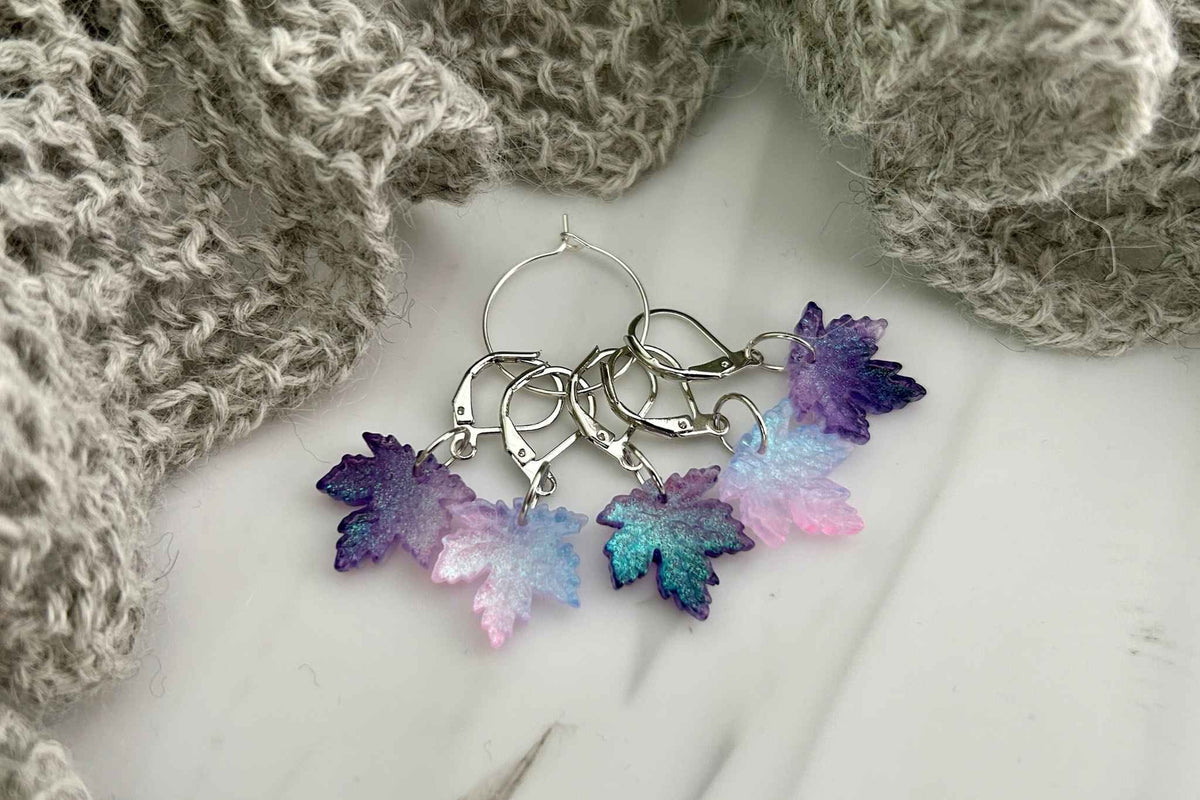 A set of 5 maple leaf knitting or crochet stitch markers or progress keepers with dangling maple leaf charms in purples, blues, and light pinks, displayed on a white marble surface with a gray knitted scarf.