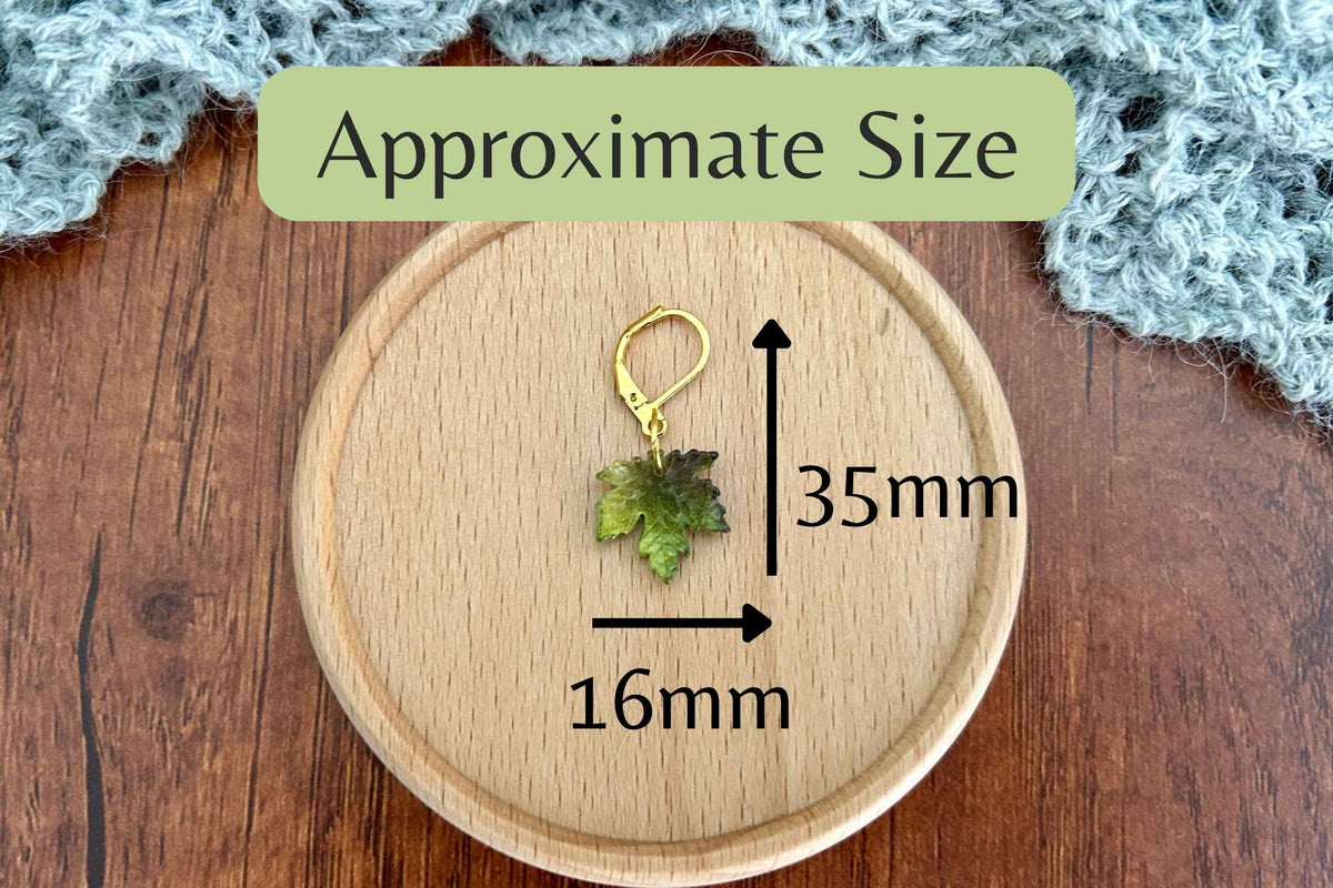 A small leaf-shaped progress keeper or stitch marker with a gold clasp is shown on a round wooden tray, with overlaid arrows and text indicating approximate dimensions of 35mm by 16mm. The scene is styled on a wooden surface with a soft gray textile in the background.