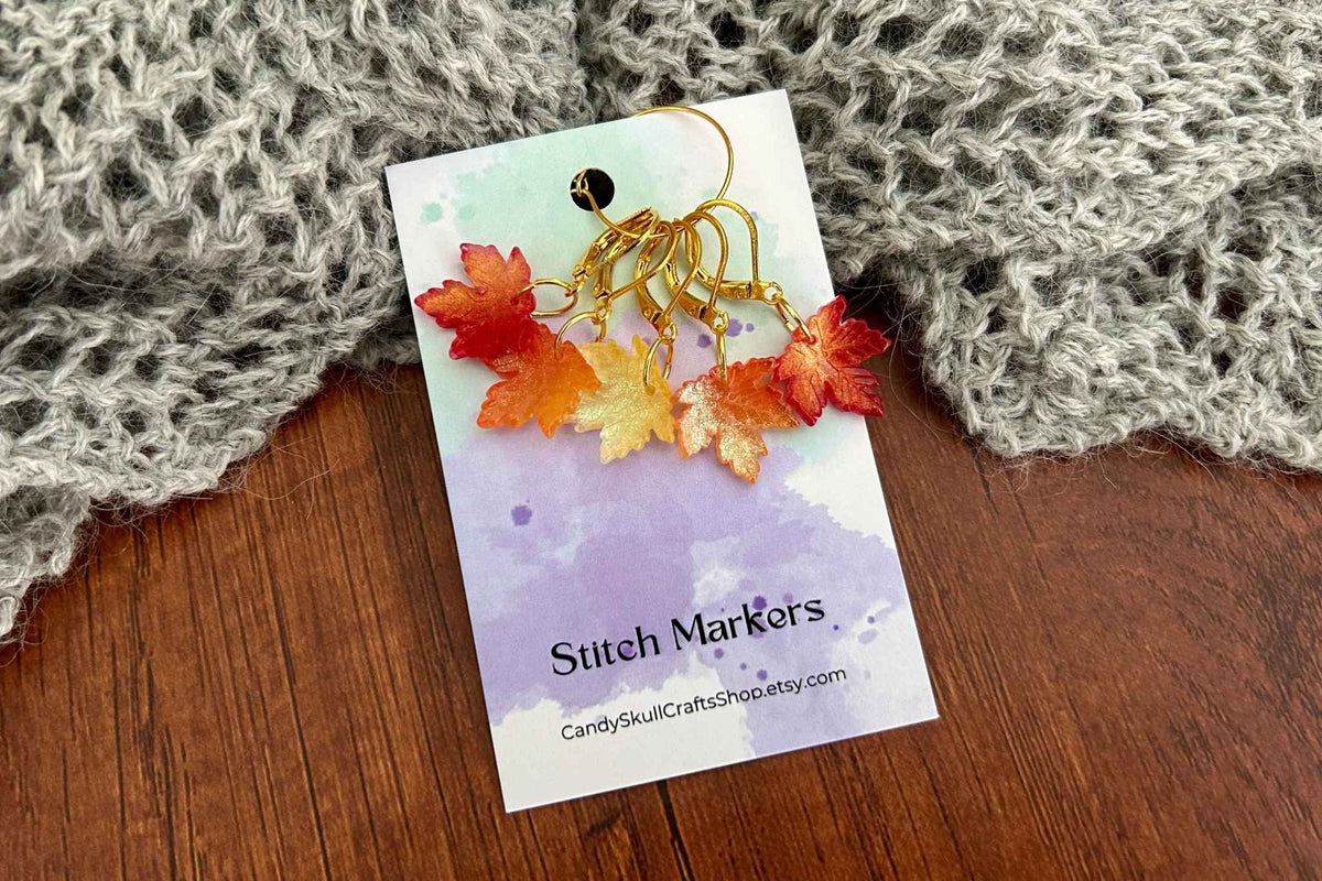 Autumn leaf stitch markers on a branded display card, styled on a wooden surface with a gray knitted blanket in the background.