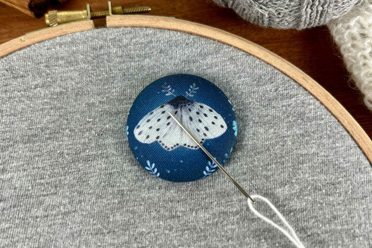 A close-up of a round fabric-covered magnetic needle minder with blue and white moth motif shown with a needle and white thread on a gray fabric background.