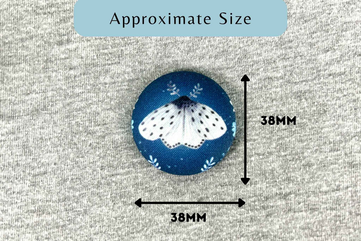 A round fabric magnetic needle minder with a blue background and a white moth design, shown with measurement arrows and text indicating approximate size 38mm by 38mm on a gray textured fabric background.