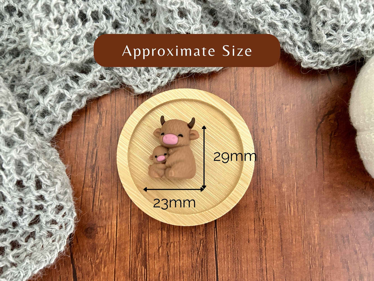A single highland cow stitch saver, overlaid with dimension labels reading 23mm and 29mm and a brown banner that says Approximate Size. The needle stopper is placed on a wooden surface with a gray knitted textile in the background.