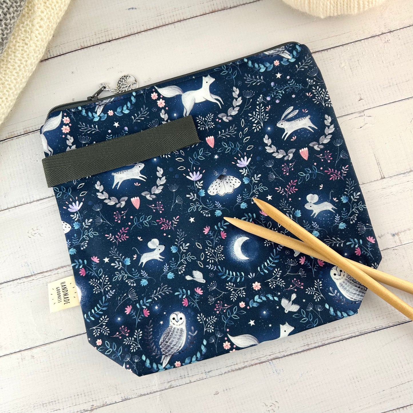 A dark blue knitting or crochet project bag with grey handle & zip laid flat on a wooden surface. The fabric features nocturnal woodland animals - a hare, fox, moth, and owl