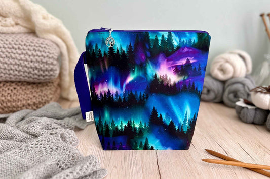 A fabric zippered knitting or crochet project bag with a colourful aurora borealis and forest pattern, standing on a wooden surface surrounded by cosy knitted blankets and yarn.