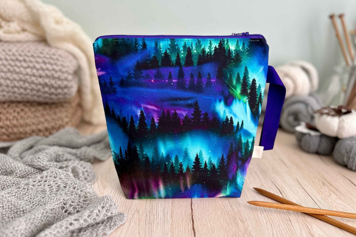 A fabric zippered knitting or crochet project bag with a colourful northern lights and pine forest print, styled on a wooden table with cosy knitted blankets and knitting tools in the background.