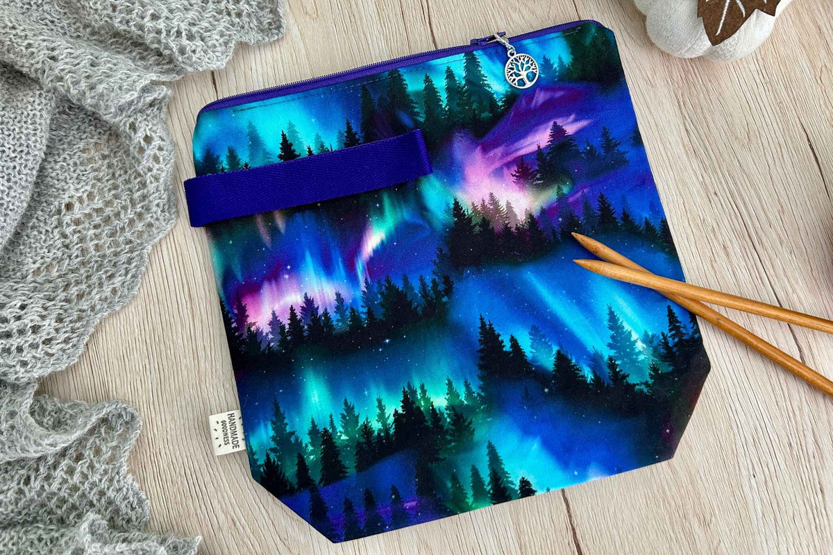 A fabric knitting or crochet project bag with a colourful aurora borealis forest print, featuring a purple handle and zip, styled on a light wood background with knitting needles and a gray knit blanket.